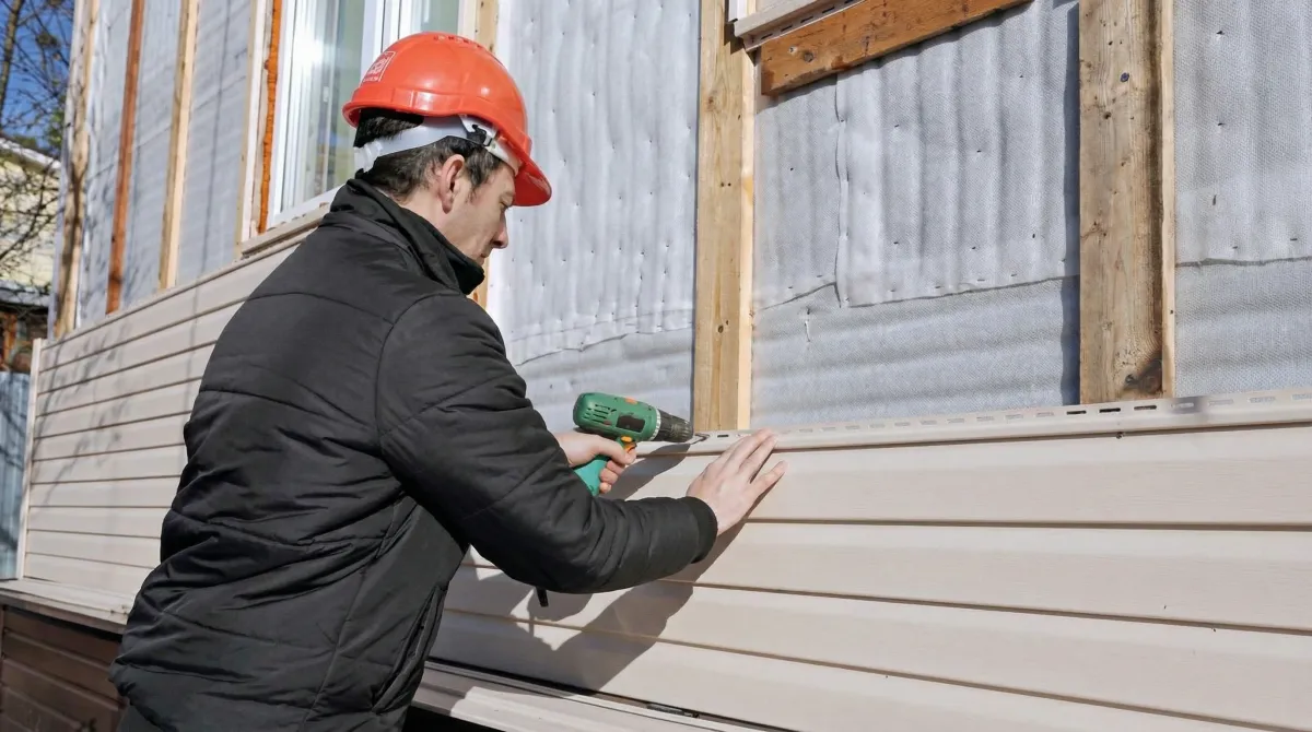 Siding replacement professional using a power drill to install new tan vinyl siding on a home exterior in Boston, MA